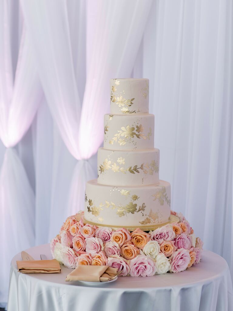 glam four tier wedding cake with gold metallic leaf design atop a bed of blush and orange roses