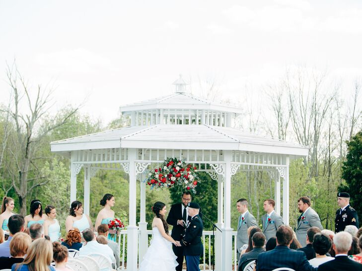 The couple exchanged vows outdoors beneath a grand gazebo structure decorated with bold red and white floral designs.