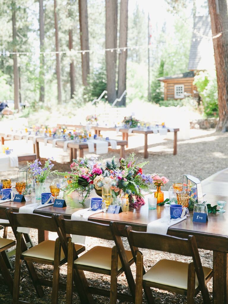 outdoor wedding reception with dark wood banquet table decorated with colorful low centerpiece and blue and yellow glassware