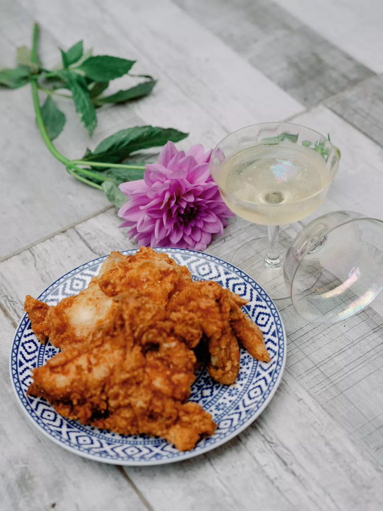 A plate of mouthwatering fried chicken sits beside a purple dahlia and a vintage punch glass.