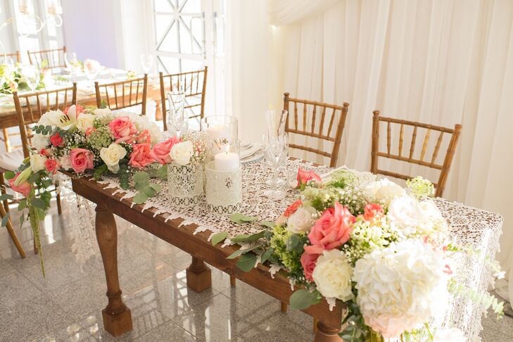 Natalie and Chris's sweetheart table had a lace runner that channeled their vintage decor inspiration. Two arrangements of white hydrangeas, baby's breath and ivory and coral roses with pops of greenery draped over the table ends.