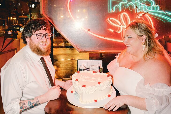 A bride and groom at their elopement party at a local college diner in Richmond, Virginia, with a heart-shaped retro-inspired wedding cake with "technically married" and a replica of an insurance card to funnily honor their logical reason for eloping.