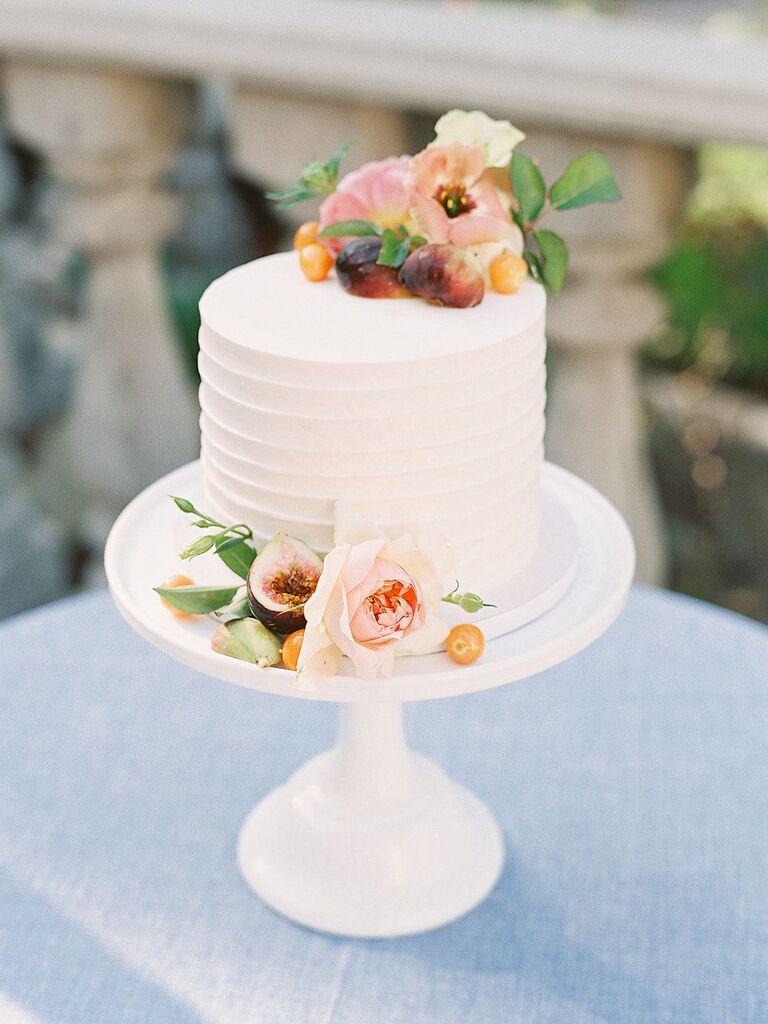 Simple one-tier wedding cake with fresh fruit and flowers atop white cake stand