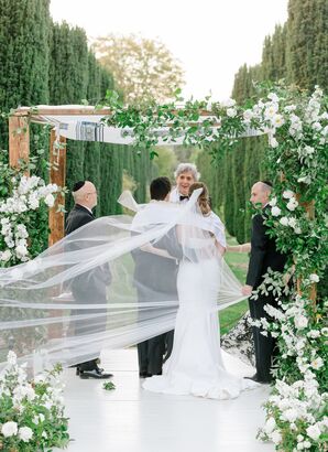 Couple Standing Under Greenery-Covered Chuppah With Veil Flowing in the Wind