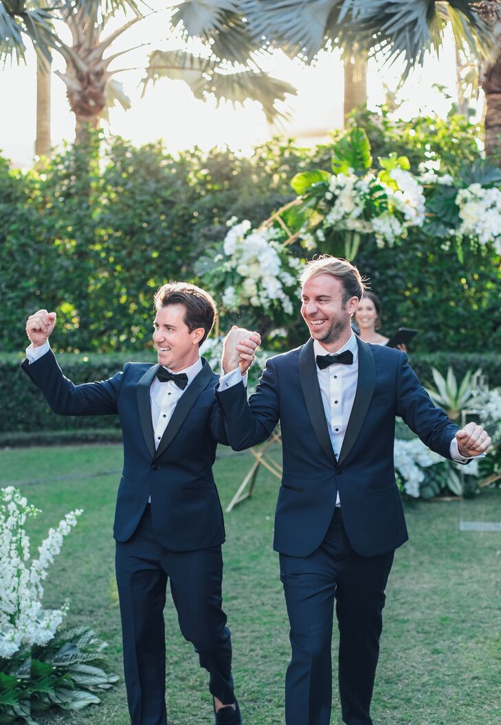 Grooms in Matching Tuxedos Holding Hands, Walking in Recessional From Green Outdoor Wedding