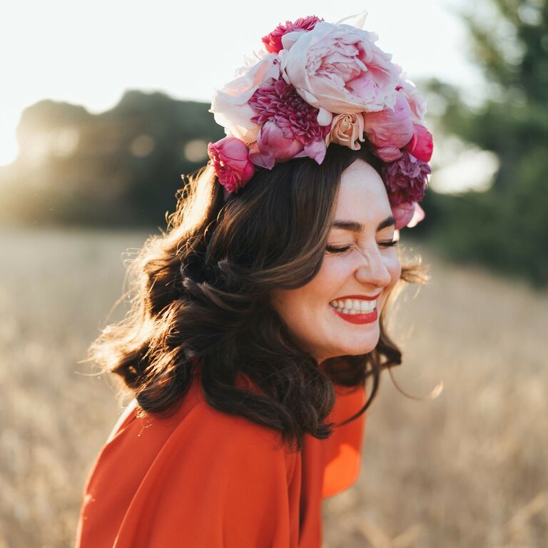 Woman wears a vibrant pink floral headpiece while laughing in a field. 