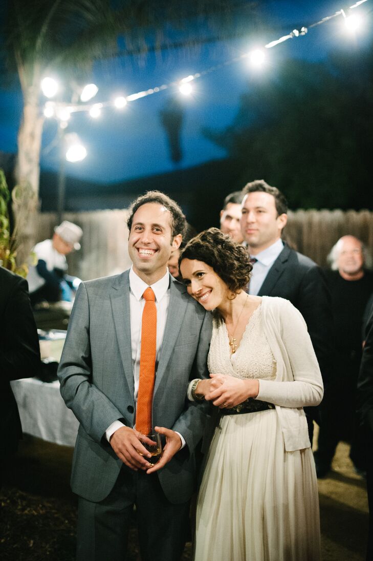 The newly married couple leaned on one another underneath twinkling lights during the backyard reception. Chris changed his tie from black to orange for the reception, and Emily put on a cardigan over her ivory wedding dress.