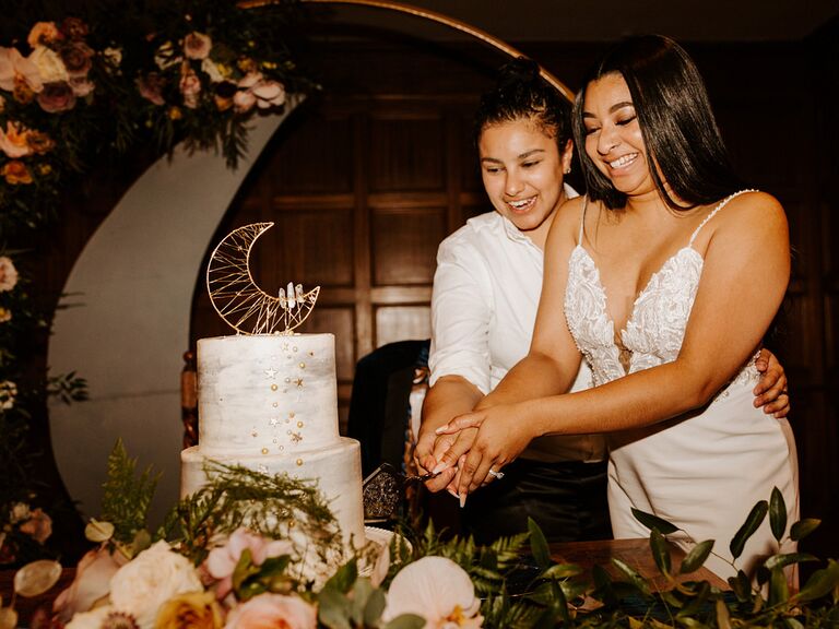 Brides cutting wedding cake with gold moon topper at celestial wedding