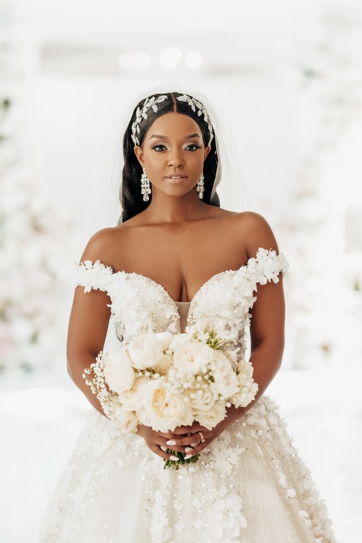 A bride in a glamorous makeup look with crystal drop earrings and ornate, large headpiece and a long veil with an off-the-shoulder wedding gown with 3D floral details and an all-white bouquet at a luxurious wedding.