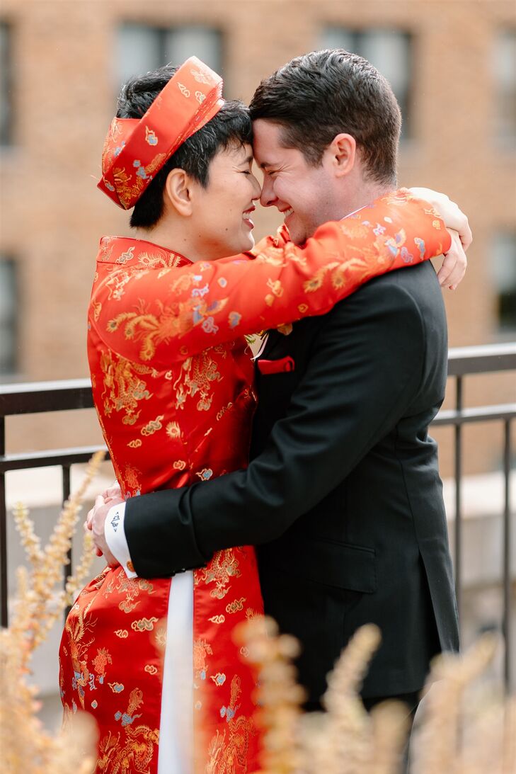 A couple in a traditional red Vietnamese ao dai outfit and a classic black suit with red details embrace on the balcony before their elegant and modern wedding with a blend of their cultural heritages.