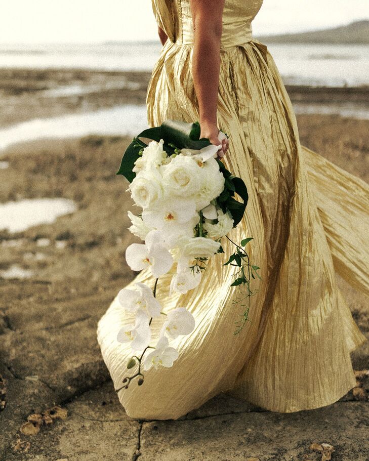 A large, cascading bouquet with ferns and greenery, as well as white orchids and roses in the hands of a bride dressed in a stunning gold-metallic dress on a city beach.