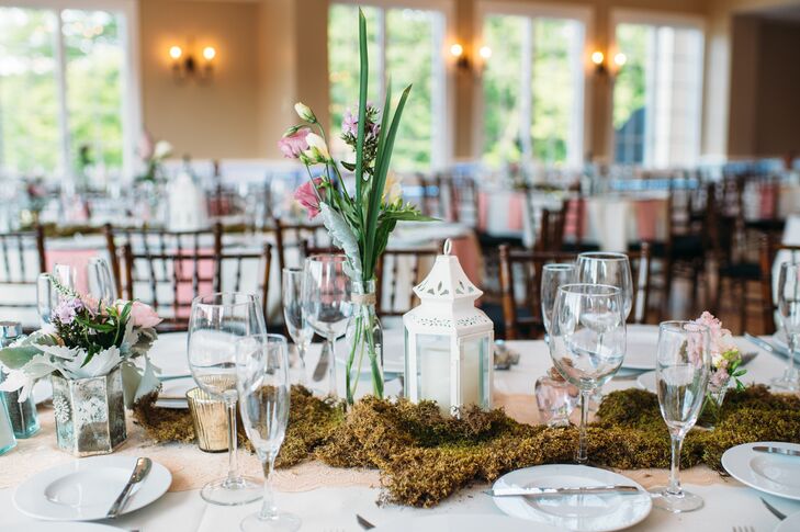But Kim and Nick ensured their oval seating stood out with added natural flair. Lines of moss filled the tablescapes alongside various tealight candles in mixed metal votives. Bulb vases filled with pink roses and greenery as well as low pink delphinium arrangements and more textured displays also highlighted the table. Champagne lace runners created a soft contrast against the ivory linens.