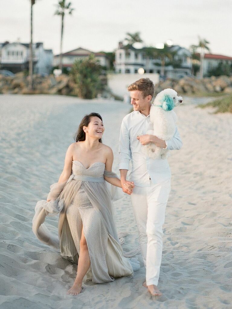 Couple holds hands and walks down the beach with their dog. 