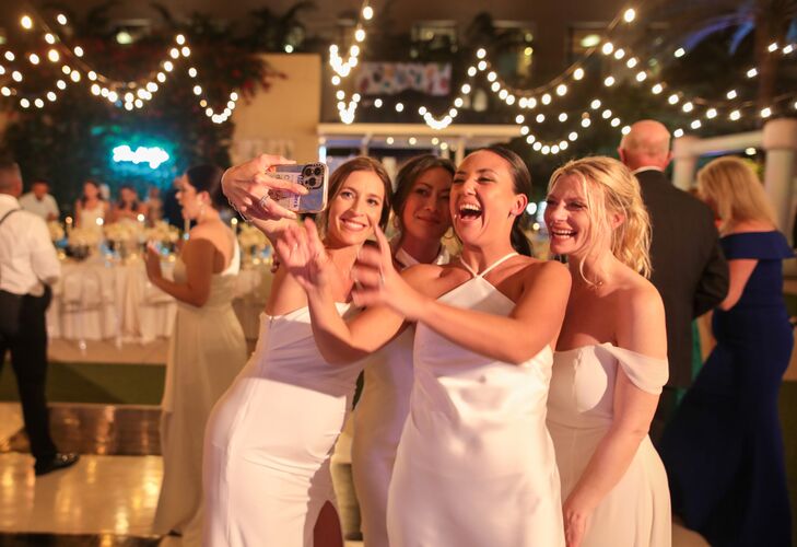 Bridesmaids in White Dresses Taking Selfies on the Dance Floor Under String Lights, Reception