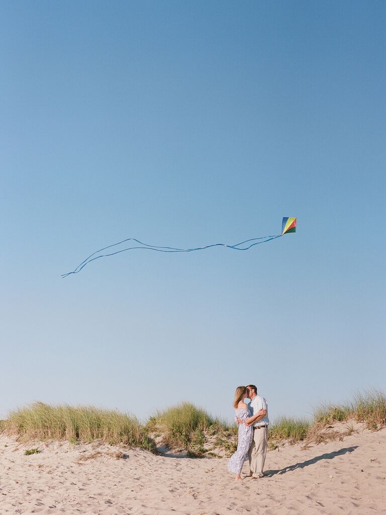 Couple embraces on a beach while a kite flies above them. 