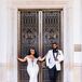 A bride in a fitted bridal gown with a deep v-neckline bedecked with crystals and off-the-shoulder straps sporting a glamorous, Old Hollywood down hairstyle, matte makeup and a crystal headpiece poses outside of the Baltimore Museum of Art with her groom in a white tuxedo jacket with black trim and black slacks before their black-tie wedding.