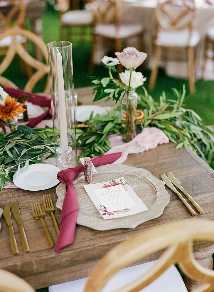 A fall, bohemian place setting on a long wooden table featuring a charger plate, gold flatware, a floral-themed menu, deep burgundy napkins, a greenery runner, long candles and low floral centerpieces.