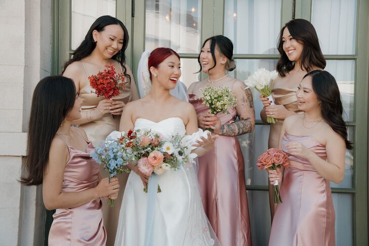 A bride in a sleek A-line gown with detached sleeves and a large bouquet of different pastel flowers posing with her bridesmaids in different satin dresses, all holding varying bouquets of single-flower bunches at a historic venue in Los Angeles.