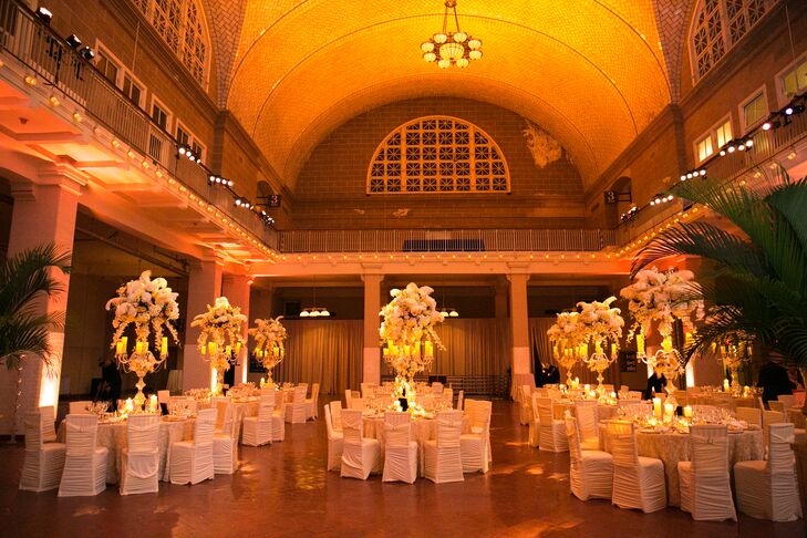 As guests made their way into the Registry Room for the reception at Ellis Island in New York City, they were immediately blown away by the transformation. Warm uplighting and skyward-reaching arrangements of full blooms introduced an element of drama to the historic room, while soft candlelight created a feel of undeniable romantic ambiance. Gilded dinnerware, crystal accents and rose-tufted cream linens had a luxe vibe that achieved the glamorous, old-Hollywood feel the couple were after.