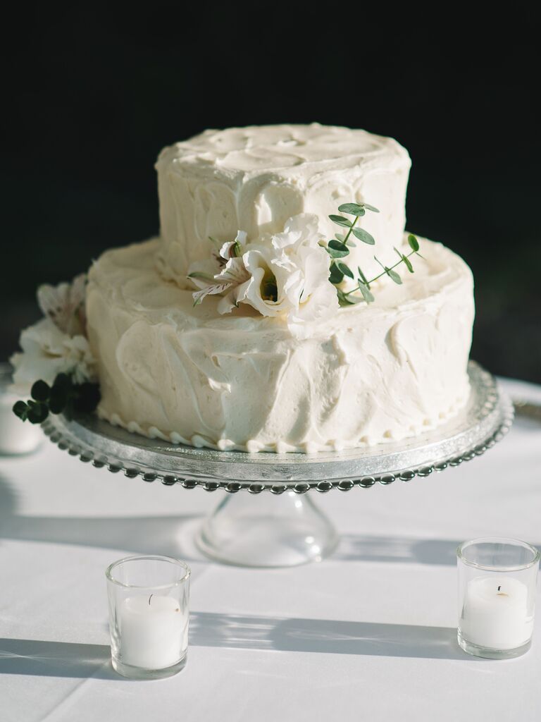 simple two tier wedding cake with textured buttercream and white flowers on top of glass wedding cake stand