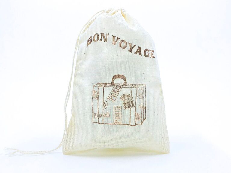 White drawstring bag with 'Bon Voyage' embroidered in sans-serif brown type above embroidered luggage icon