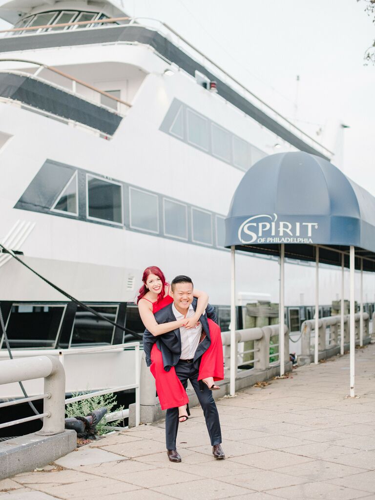 Wife poses on her husbands back with a large boat in the background. 