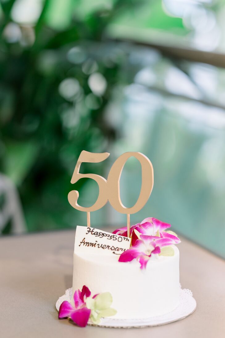 A small, white, single-tier cake decorated with striking pink orchids with fondant and a gold cake topper with "50" for the couple's 50th wedding anniversary and vow renewal in Hawaii.