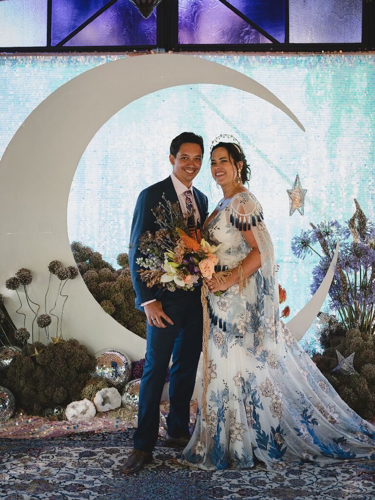 Bride and groom posing in front of crescent moon backdrop at celestial wedding
