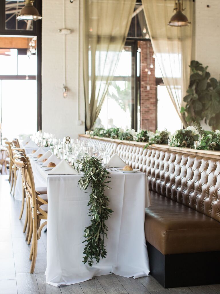 long wedding banquet table with white tablecloth, greenery table runner and wooden cross-back chairs