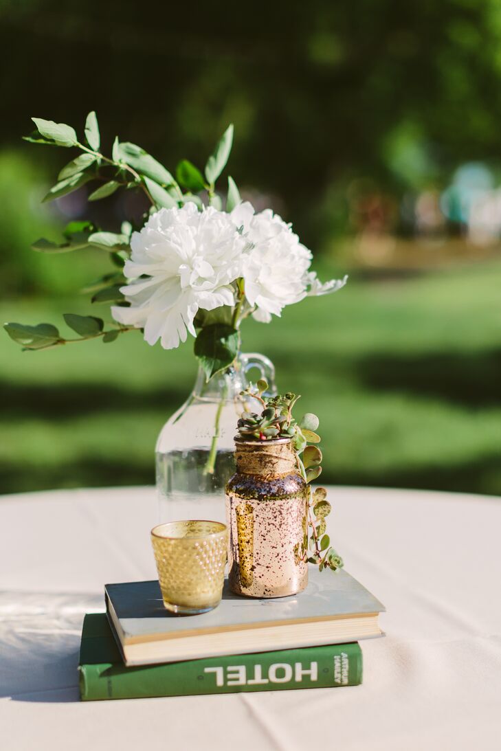 Ivory chrysanthemums and green leaves arranged inside glass jars sat on top of a vintage stack of books, positioned next to another vase and votive candle.