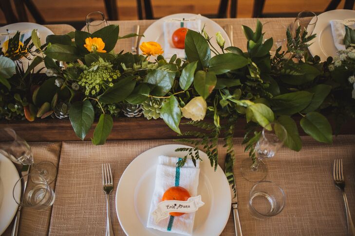 Dining tables were covered in burlap linens and set with white dinnerware. The white-and-blue-striped napkins had a pieces of fresh fruit and white seating assignments on top for guests to find their spot at the tables. Lush arrangements of leaves, succulents and flowers filled rustic wooden boxes that served as dining table centerpieces.