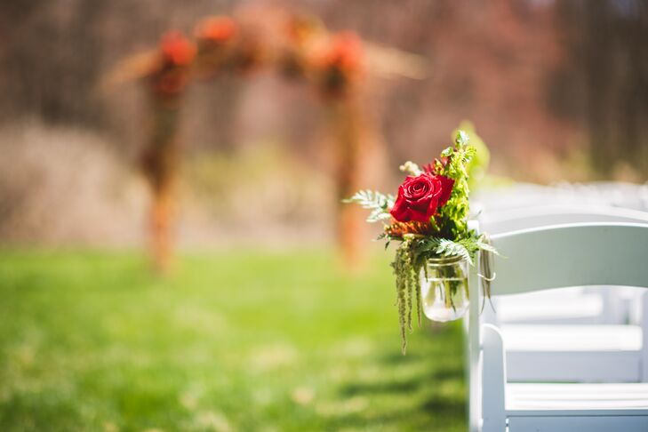The same flowers picked for their centerpieces, bouquets and boutonnieres were used in the aisle decor. Lush arrangements of red roses, orange proteas and greenery were placed in mason jars and hung against each row of white folding chairs.