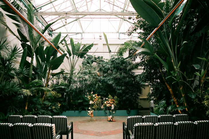 A verdant greenhouse ceremony space with an industrial feel featuring towering and low plants and trees, unique ceremony chairs and a low floral altar arch with burnt orange and white flowers at an edgy, trendy wedding in Downtown LA.