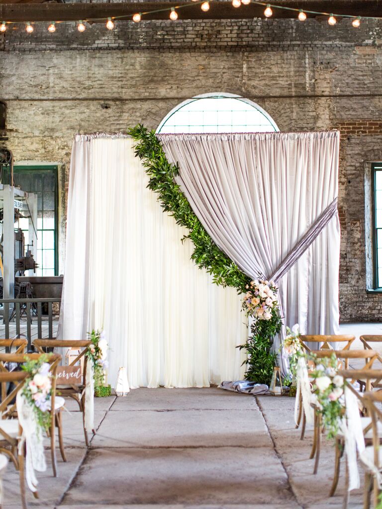indoor wedding ceremony backdrop with curtain draped to one side decorated with greenery garland and flower tie-back
