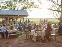 outdoor rehearsal dinner venue with guests seated at round tables with cross. back wooden chairs and string lights hanging above