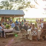 outdoor rehearsal dinner venue with guests seated at round tables with cross. back wooden chairs and string lights hanging above