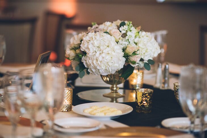Some of their reception tables also featured low, garden-style arrangements. White hydrangeas, blush garden roses, seeded eucalyptus and scabiosa pods flowed along the sides of its gold pedestal vase. A mirror base elevated the design. Gold votive candles also surrounded the arrangement.