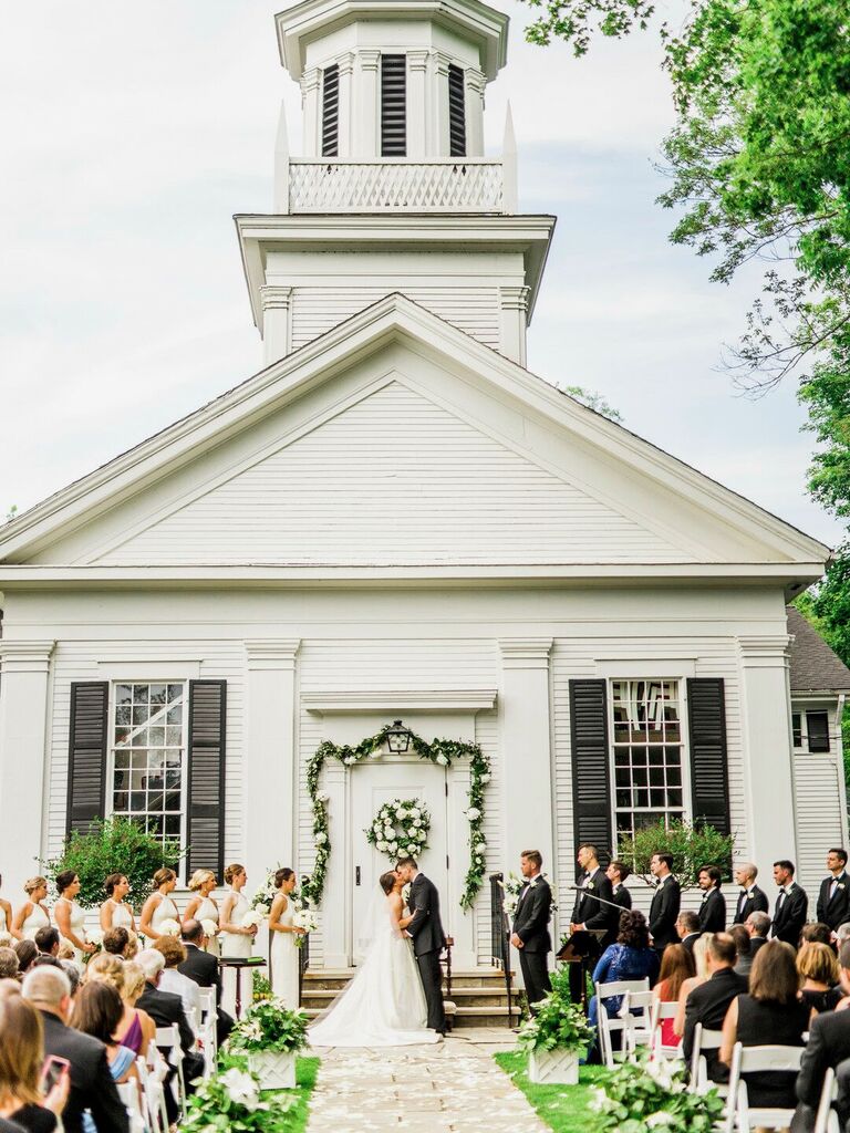 bride and groom kiss in front of white wedding chapel door decorated with greenery garland