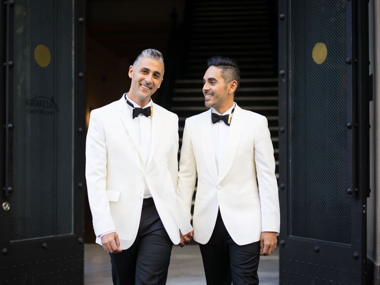 two grooms holding hands while exiting a building and wearing matching white tuxedo jackets with black bow ties