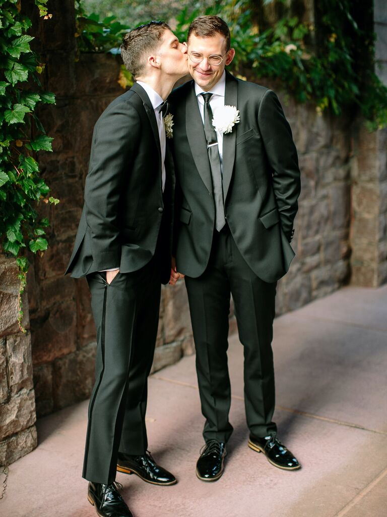 two grooms standing in garden wearing black tuxedos one groom kisses the other's cheek
