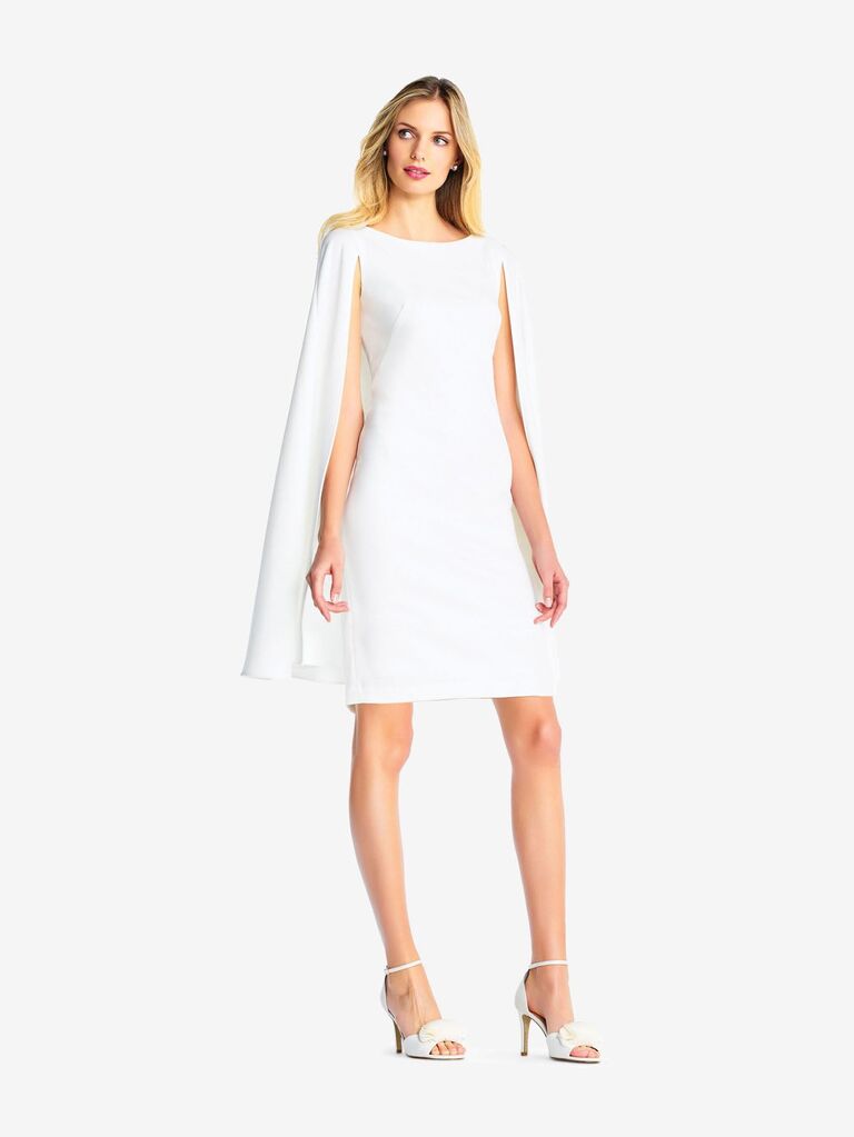 Model wears a short white dress with an attached cape. 
