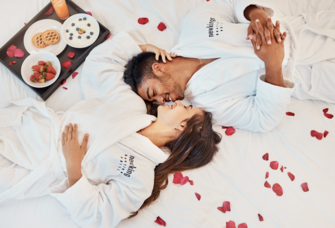 Couple laying on hotel bed in bath robes surrounded by rose petals and breakfast
