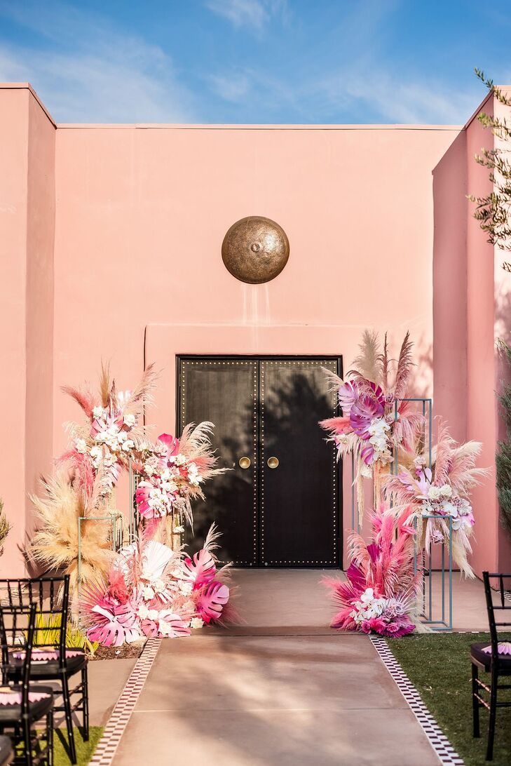 A beautiful, mid-century modern wedding ceremony space with tons of pink, fuchsia and blush details--featuring pampas grass, painted palm leaves and flowers set against a pink wall backdrop at the famous The Sands Hotel in the California desert in Indian Wells/Palm Springs.