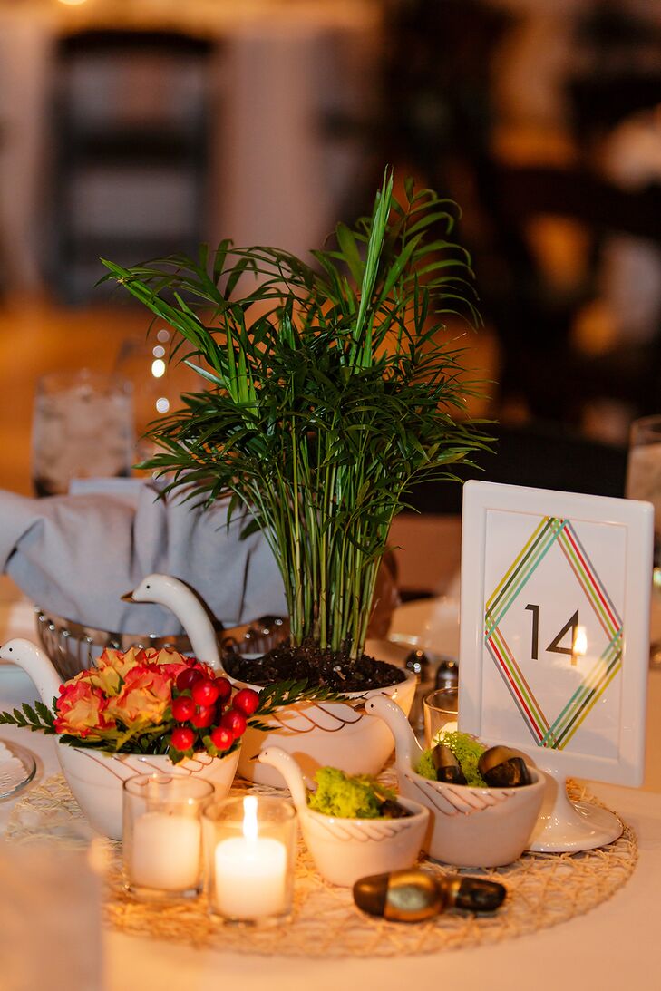 Neanthe bella palm plants, mambo spray roses and red hypericum berries with leather leaf fern and groupings of reindeer moss with gold painted rocks were each nestled in a ceramic measuring cup. White table numbers had the evening's menu printed on the other side.