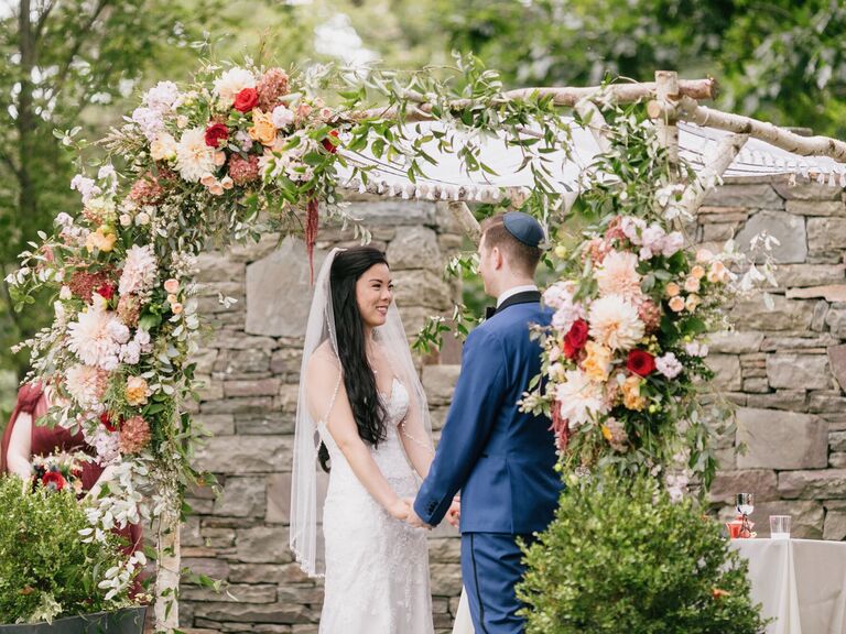 bride and groom standing under chuppah made from birch tree branches decorated with flowers and greenery