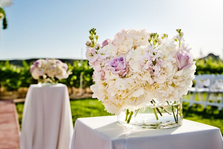 The aisle was lined with white hydrangeas, purple roses and stock arranged in glass vases, positioned on top of tall tables covered in white linens.