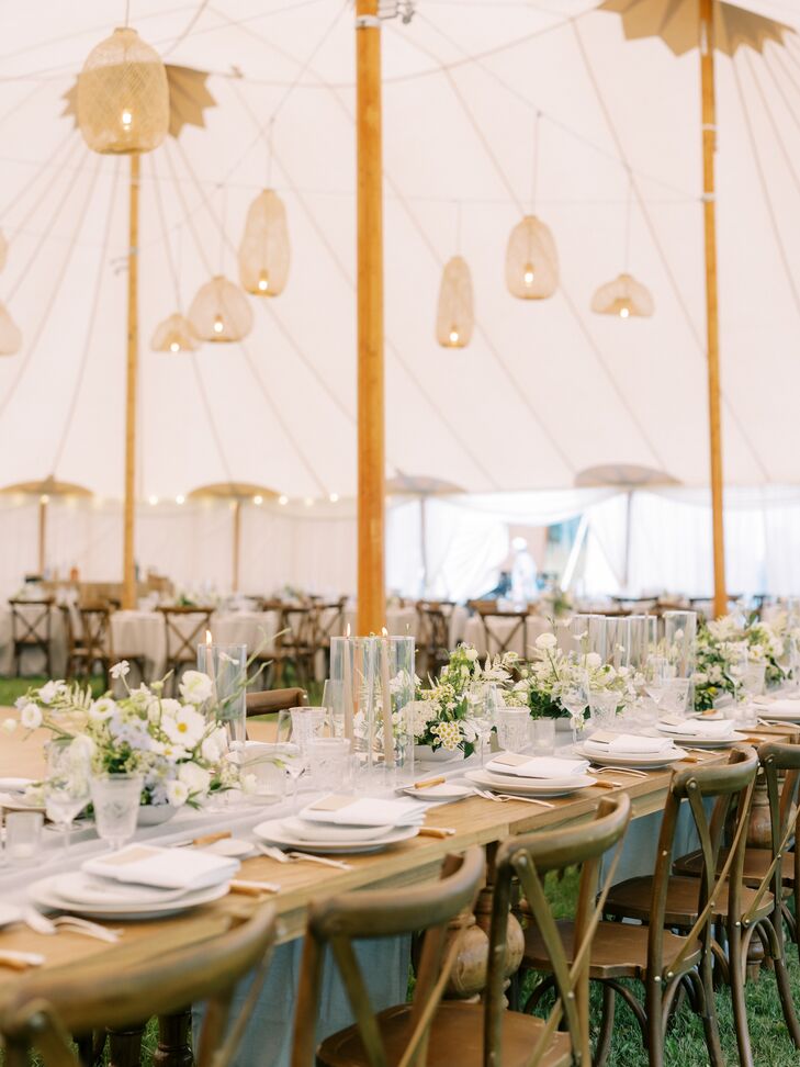 A rustic, tented wedding reception with rattan chandeliers, long and round tables, low centerpieces with white flowers and glass-encased candles.