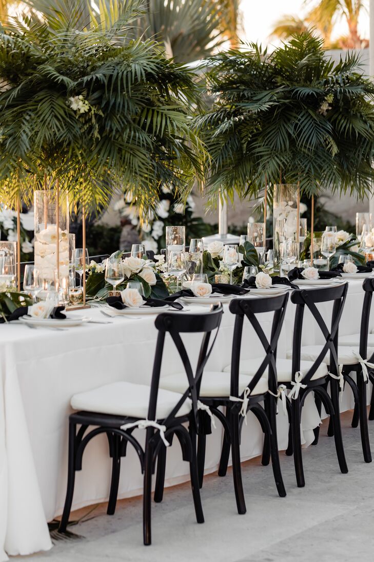 A long table with white table linens, tall and short centerpieces featuring tropical greenery, low and large candles, a floral table runner made up of greenery, and black-and-white details.