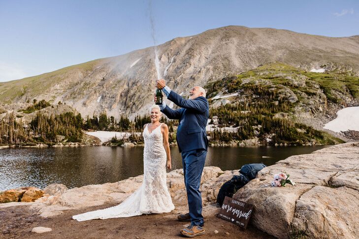 A groom in a navy suit pops a bottle of Champagne as his bride in a fitted wedding gown watches at their lakeside Rocky Mountain elopement.