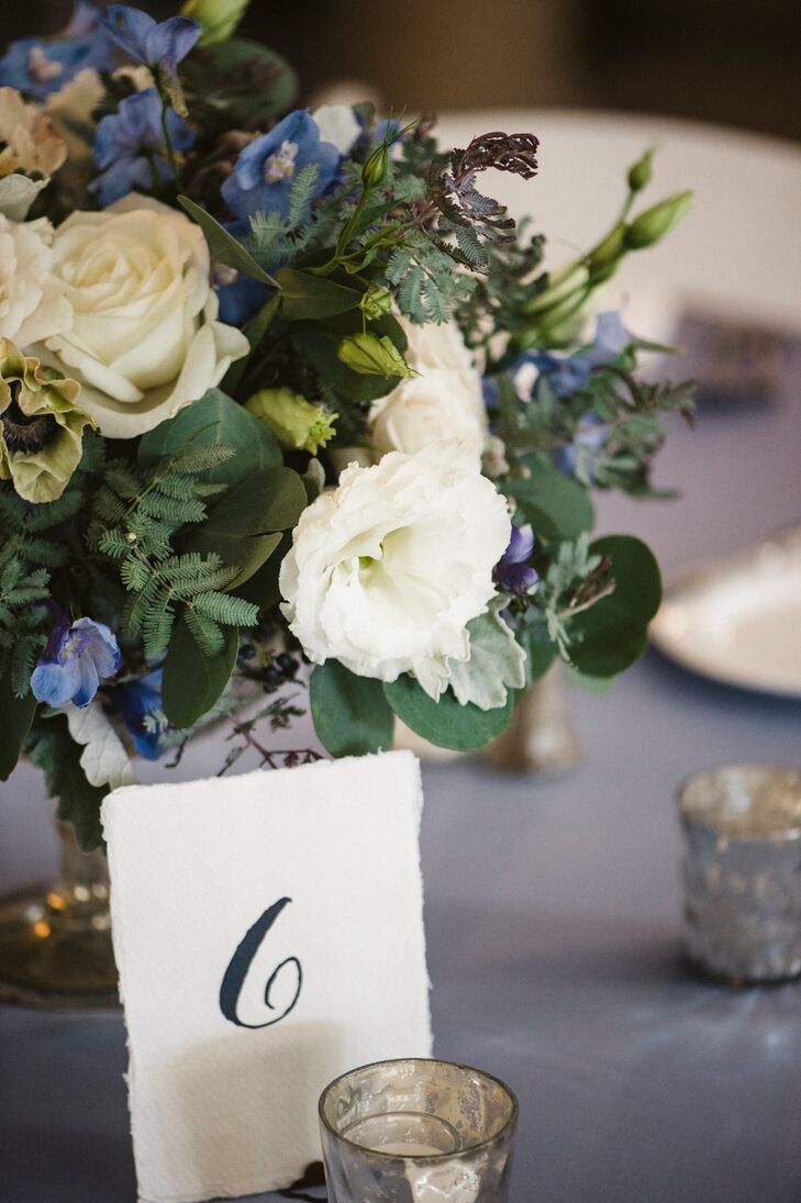 One of centerpieces was a blue and white floral arrangement, including delphinium in varying shades, white miniature spray roses, white lisianthus, blue viburnum berries, silver brunia berries, white anemones and white roses accented with dusty miller and seeded eucalyptus foliages, with five silver mercury-glass votive candles placed around the vase.